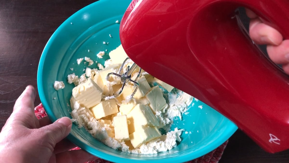 A person uses a red electric hand mixer to blend cubed butter and powdered ingredients in a turquoise mixing bowl while preparing an easy strawberry pound cake.