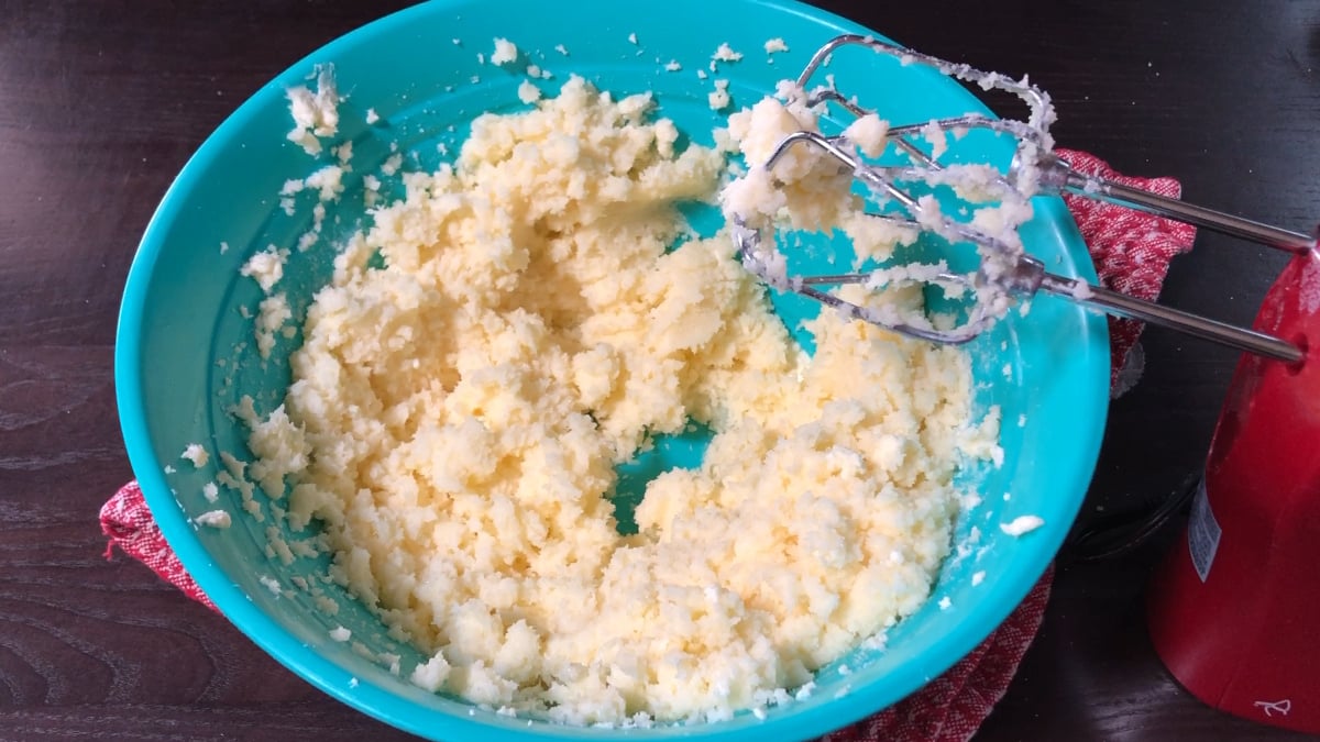 A blue bowl with creamed butter and sugar sits on a red towel, ready to become a moist strawberry pound cake, next to a hand mixer with some mixture clinging to the beaters.