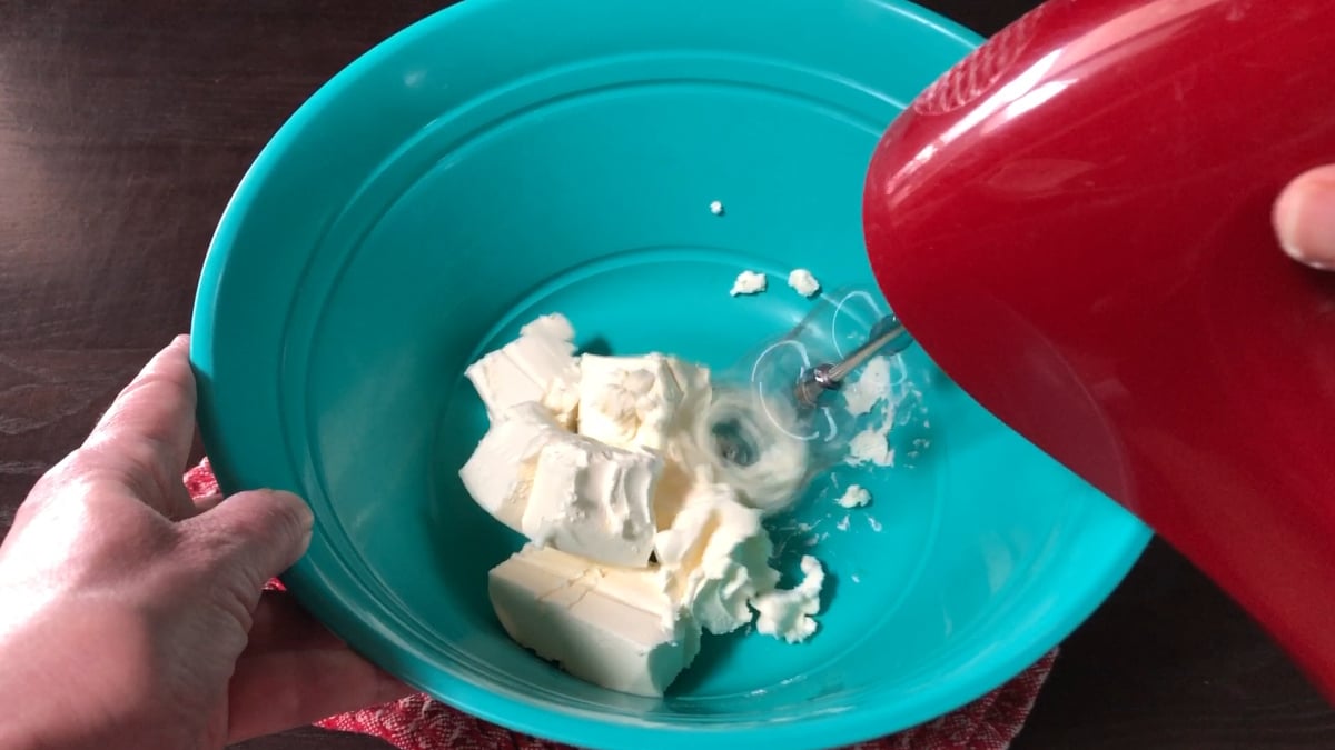 A person uses a red hand mixer to blend cream cheese in a teal mixing bowl on a table, preparing ingredients for a moist strawberry pound cake.