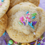A close-up of soft sugar cookies topped with colorful sprinkles, with extra sprinkles scattered on a white surface below.