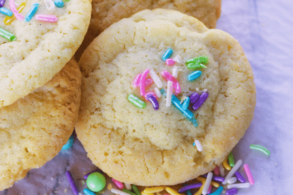 A close-up of soft sugar cookies topped with colorful sprinkles, with extra sprinkles scattered on a white surface below.