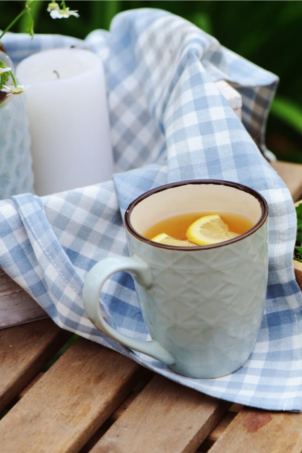 A blue mug of tea with lemon slices rests on a blue checkered cloth next to an unlit white candle on a wooden surface, embodying the essence of simple living and minimalism.