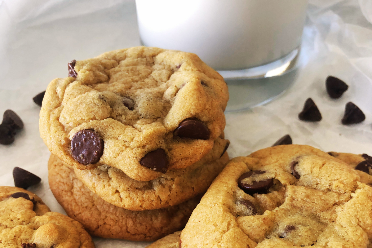 Stack of chocolate chip cookies next to a glass of milk, with scattered chocolate chips on a white surface—these treats look just like they were made from the classic McDonald’s chocolate chip cookie recipe.