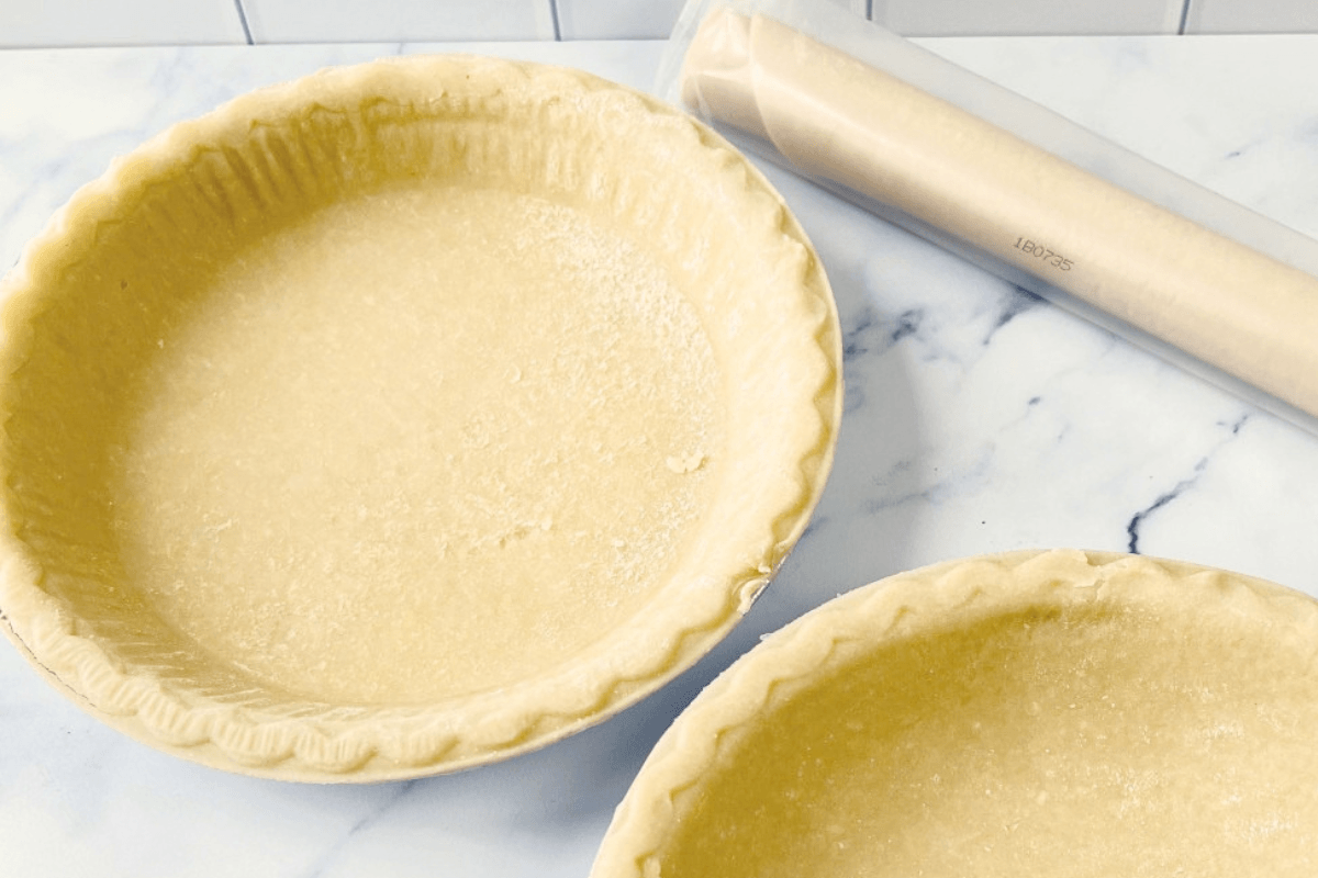 Two unbaked pie crusts in pie pans are on a marble surface, with a rolled-up sheet of parchment paper nearby—perfect for learning how to use frozen pie crust or tips on thawing frozen pie crust before baking.