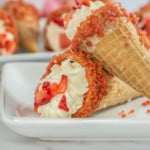 Strawberry crumble ice cream cone with whipped cream topping, lying on a white dish against a blurred dessert background.