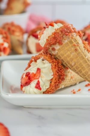 Strawberry crumble ice cream cone with whipped cream topping, lying on a white dish against a blurred dessert background.