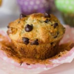 A close-up of a chocolate chip muffin partially unwrapped from its pink polka-dotted paper liner. Other muffins are blurred in the background.