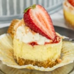 A slice of strawberry cheesecake with a whipped cream topping and a fresh strawberry half, served on a glass plate with a fork.