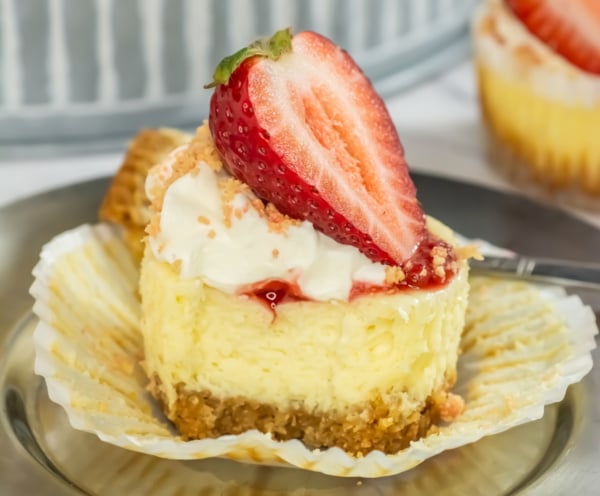 A slice of strawberry cheesecake with a whipped cream topping and a fresh strawberry half, served on a glass plate with a fork.
