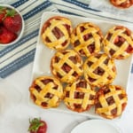 A plate of small lattice-topped strawberry pies on a striped cloth, surrounded by fresh strawberries and additional pies.