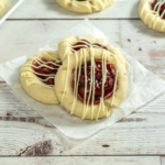 Three cherry thumbprint cookies with white chocolate drizzle rest on parchment paper on a rustic wooden surface. More cookies are visible in the background.