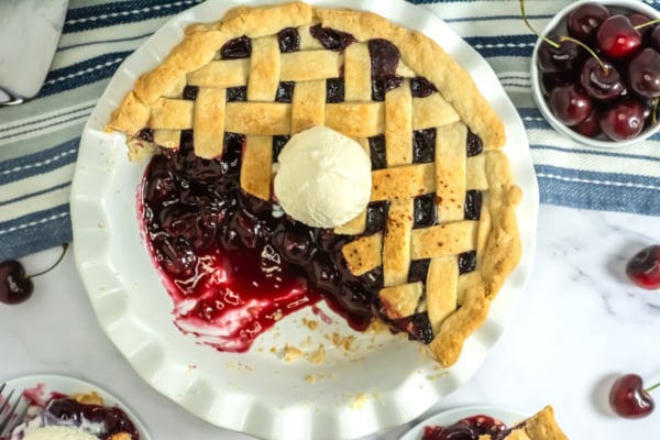 A partially eaten cherry pie with a lattice crust topped with a scoop of vanilla ice cream on a white pie dish. Fresh cherries and a striped cloth are visible in the background.