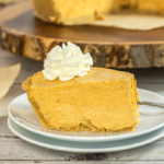 A slice of pumpkin pie topped with whipped cream on a white plate with a fork, set on a wooden table. The rest of the pie is in the background on a wooden serving board.
