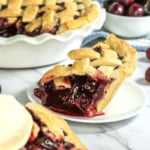 A slice of cherry pie in the foreground with a scoop of vanilla ice cream beside it. The entire pie and a bowl of cherries are in the background on a marble countertop.