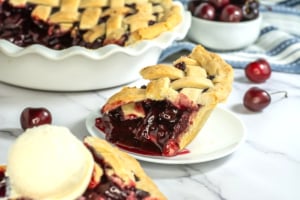 A slice of cherry pie in the foreground with a scoop of vanilla ice cream beside it. The entire pie and a bowl of cherries are in the background on a marble countertop.