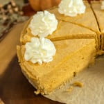 A sliced pumpkin cheesecake with dollops of whipped cream on each piece, placed on a rustic wooden board lined with parchment paper.