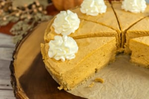 A sliced pumpkin cheesecake with dollops of whipped cream on each piece, placed on a rustic wooden board lined with parchment paper.