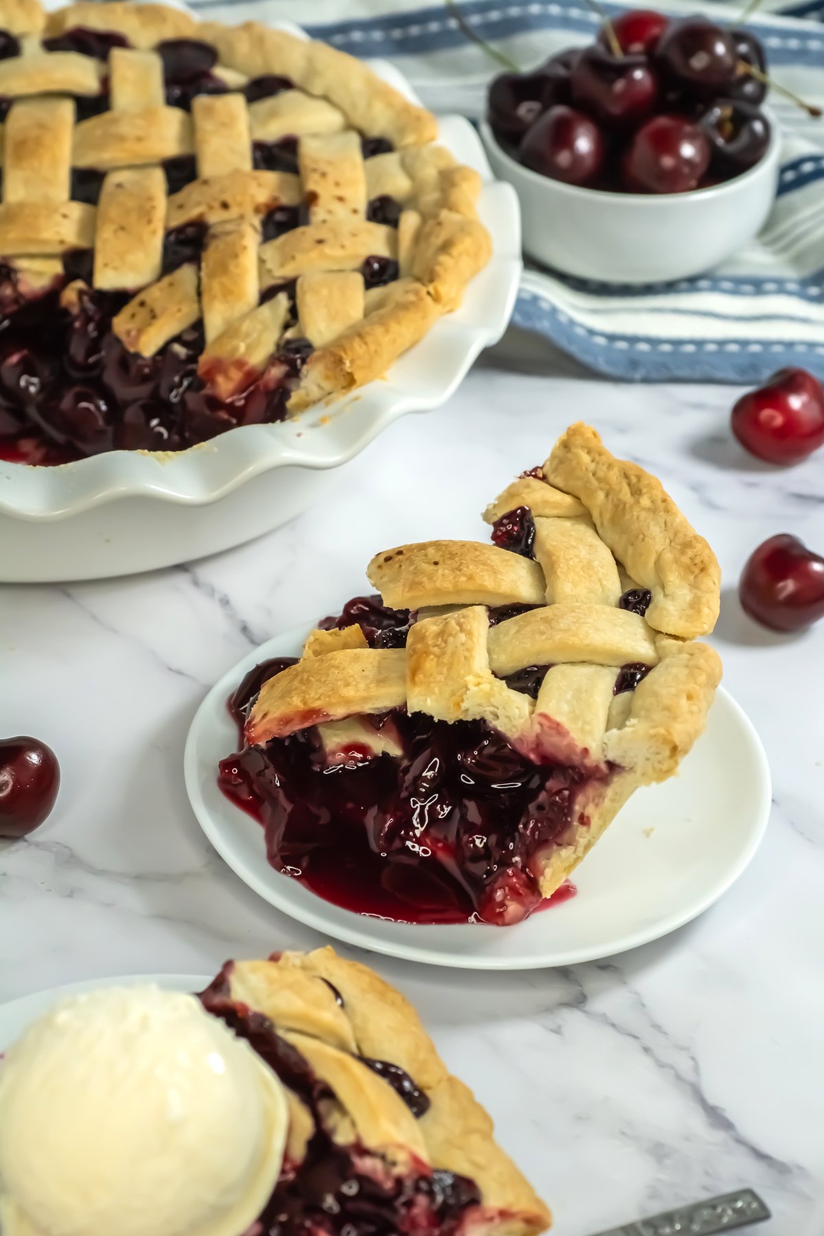 A lattice-top cherry pie with a slice served on a white plate, accompanied by a scoop of vanilla ice cream and a small bowl of fresh cherries in the background.