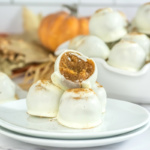 A stack of white chocolate-covered truffles on plates, with one truffle bitten to reveal a pumpkin filling, and more truffles and a pumpkin in the background.