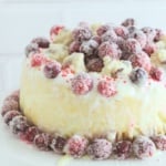 A frosted cake topped with sugared cranberries and surrounded by cranberries on a white cake stand, set against a white brick background.