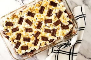 A clear glass dish filled with s'mores dessert topped with mini marshmallows, graham cracker pieces, and chocolate squares, placed next to a white and black checkered cloth.