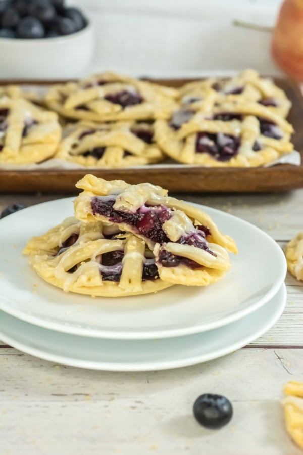 A sliced berry pastry with a lattice top sits on a stack of plates. In the background, more pastries and a bowl of blueberries are visible.