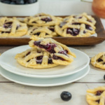 A stack of blueberry lattice pastries on plates, with more pastries and blueberries on a wooden tray in the background. An apple is partially visible.