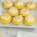 Nine lemon meringue tarts with swirled tops are arranged on a square white plate.