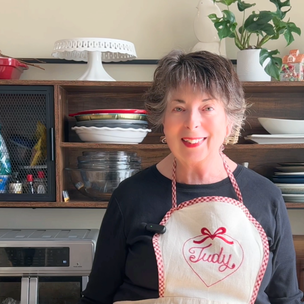 A content marketing writer with short hair, "Judy" on her apron, stands in a cozy kitchen lined with shelves of dishes and plants.