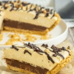 A slice of layered pie with a graham cracker crust, chocolate layer, cream filling, and chocolate drizzle, placed in front of the remaining pie in a dish.