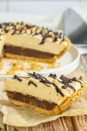 A slice of layered pie with a graham cracker crust, chocolate layer, cream filling, and chocolate drizzle, placed in front of the remaining pie in a dish.