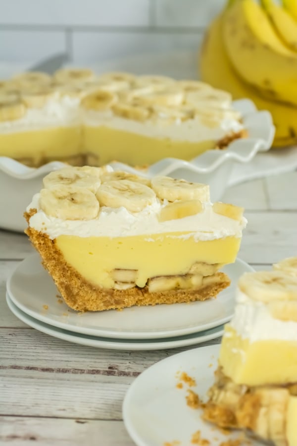 Slice of banana cream pie on a plate showing layers of banana slices, custard, and whipped cream, with a whole pie and bananas in the background.