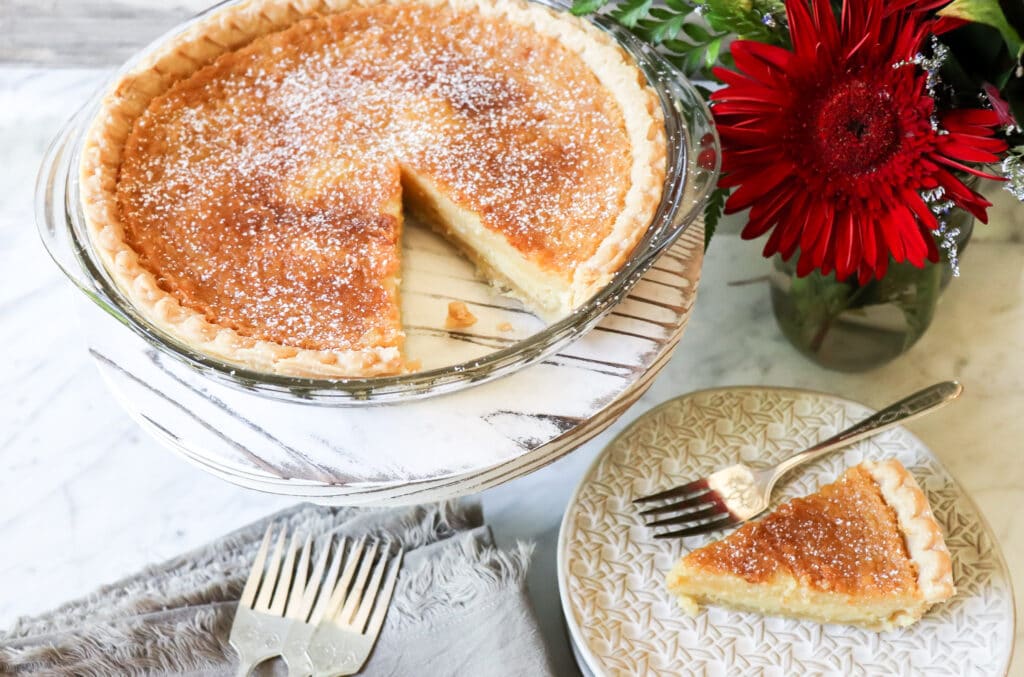 A baked pie with a slice removed sits on a stand; the slice is on a plate with a fork beside it. A red flower arrangement is next to the pie, perfect for featuring in best bloggers’ National Pie Day recipes.