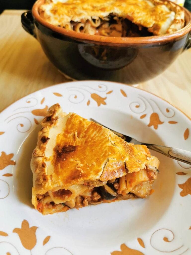 A slice of baked pasta pie with a golden crust is served on a decorative plate, perfect inspiration for easy pie recipes as you celebrate National Pie Day. The remaining pie sits invitingly in a ceramic dish in the background.