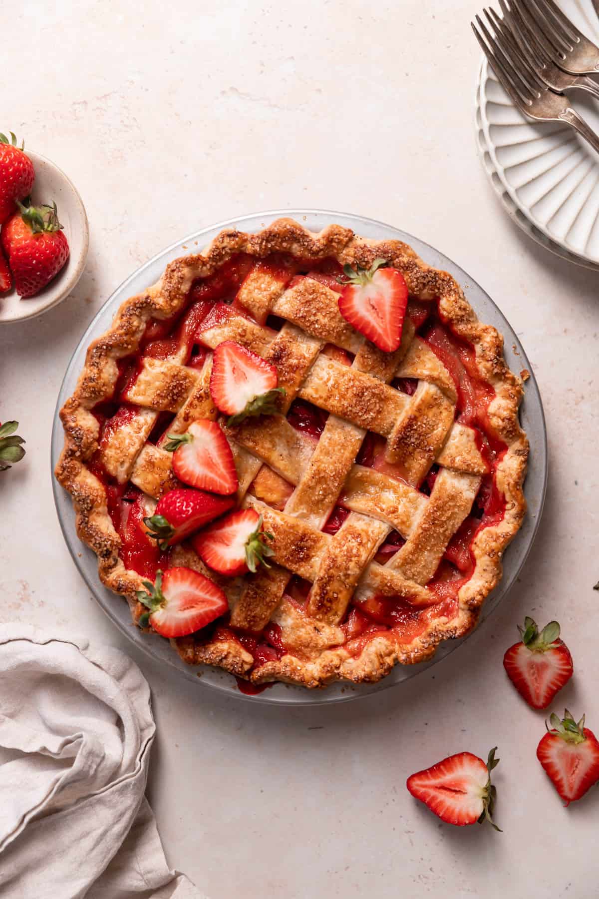 A lattice-topped strawberry pie garnished with fresh strawberry halves sits on a light surface, surrounded by plates, forks, and a bowl of strawberries—perfect for celebrating National Pie Day or adding to your list of easy pie recipes.