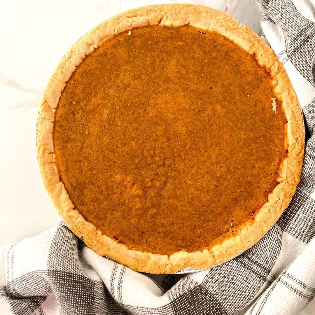 A whole pumpkin pie with a golden-brown crust, perfect for National Pie Day, sits on a white surface next to a black and white checkered cloth.