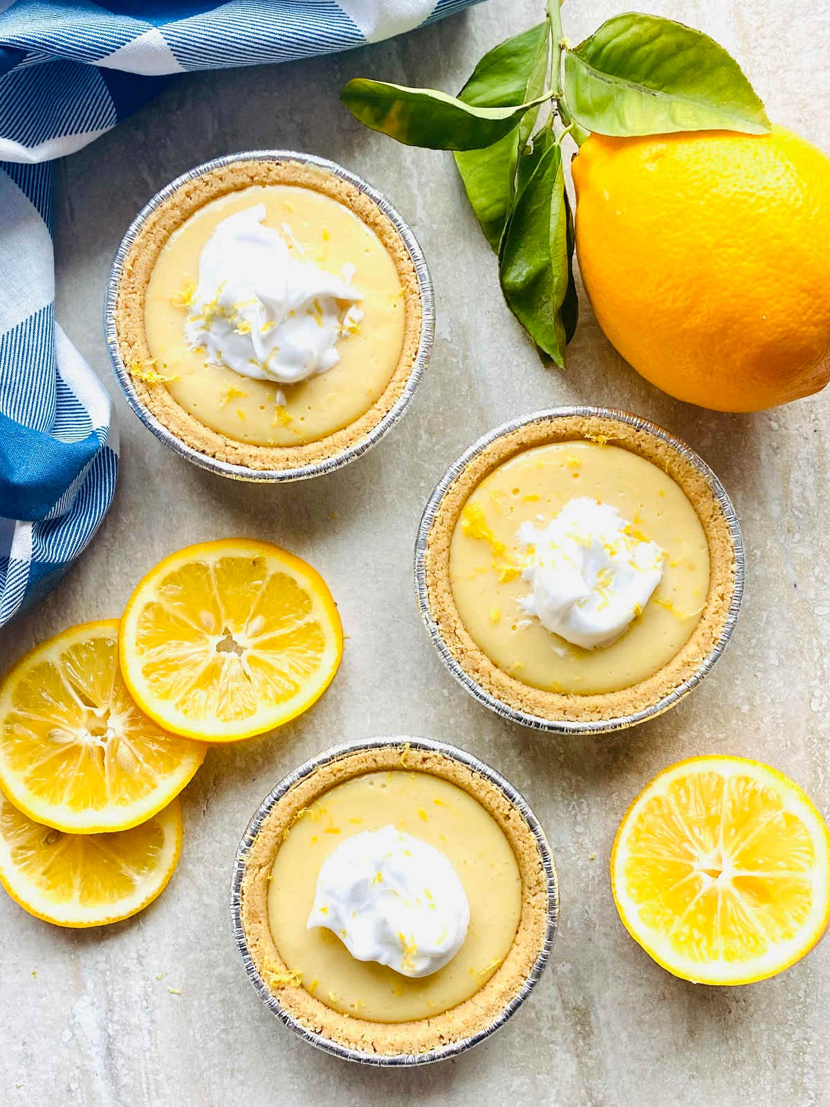 Three mini lemon pies topped with whipped cream are arranged on a light surface next to a whole lemon, lemon slices, and a blue striped cloth—perfect for National Pie Day or adding to your collection of easy pie recipes.