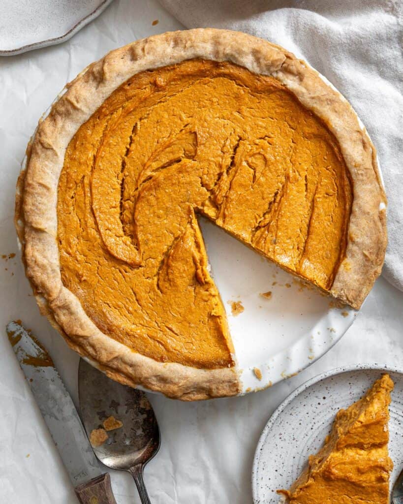 A pumpkin pie with a slice removed sits on a white surface next to a knife, spoon, and a plate holding the removed slice—perfect inspiration for easy pie recipes or celebrating National Pie Day.