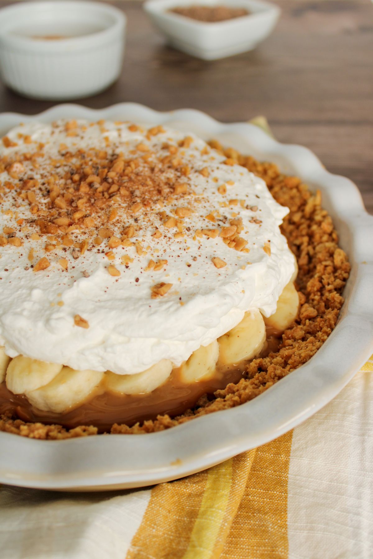 Celebrate National Pie Day with this banana cream pie featuring a crumb crust, whipped cream, and chopped nuts. Set on a yellow and white cloth, it’s one of those easy pie recipes that's sure to impress. Small bowls are in the background.