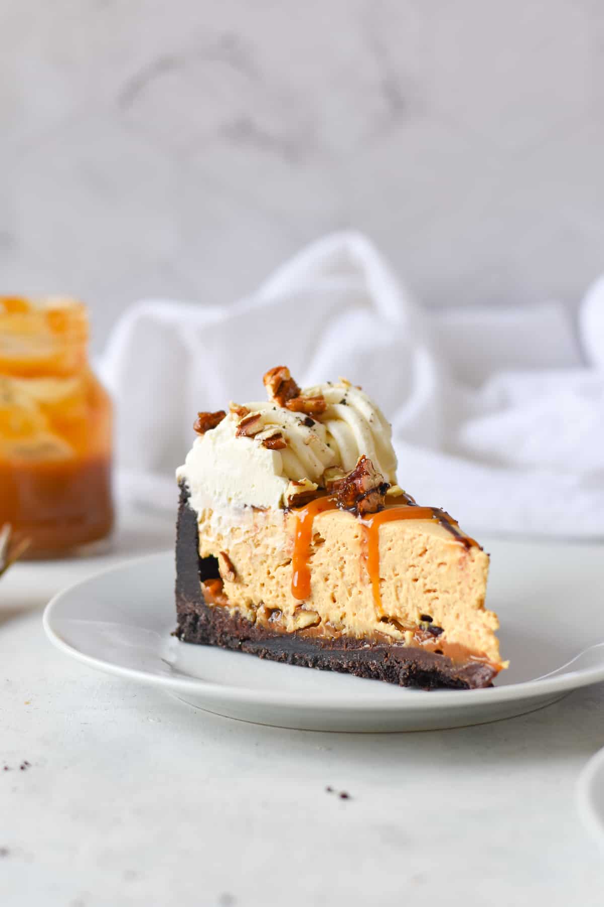 A slice of caramel cheesecake with an Oreo crust, whipped cream, caramel drizzle, and chopped nuts on a white plate—perfect inspiration for National Pie Day recipes from the best bloggers.