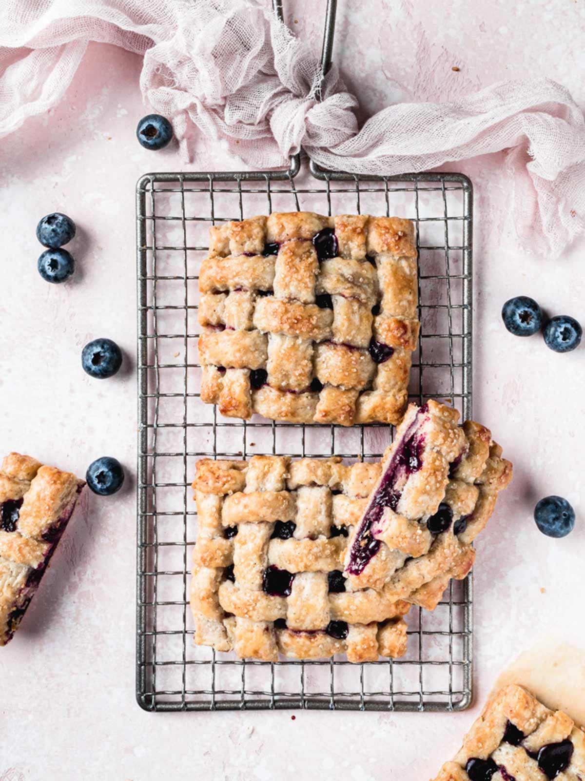 Three rectangular blueberry lattice bars rest on a cooling rack, surrounded by fresh blueberries on a light pink surface. Perfect for National Pie Day recipes, one bar is cut open to reveal the juicy filling.