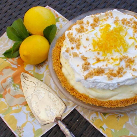 A lemon cream pie with whipped topping and lemon zest sits on a table next to two whole lemons and a pie server on a floral napkin, making it a perfect treat for National Pie Day or anyone seeking easy pie recipes.