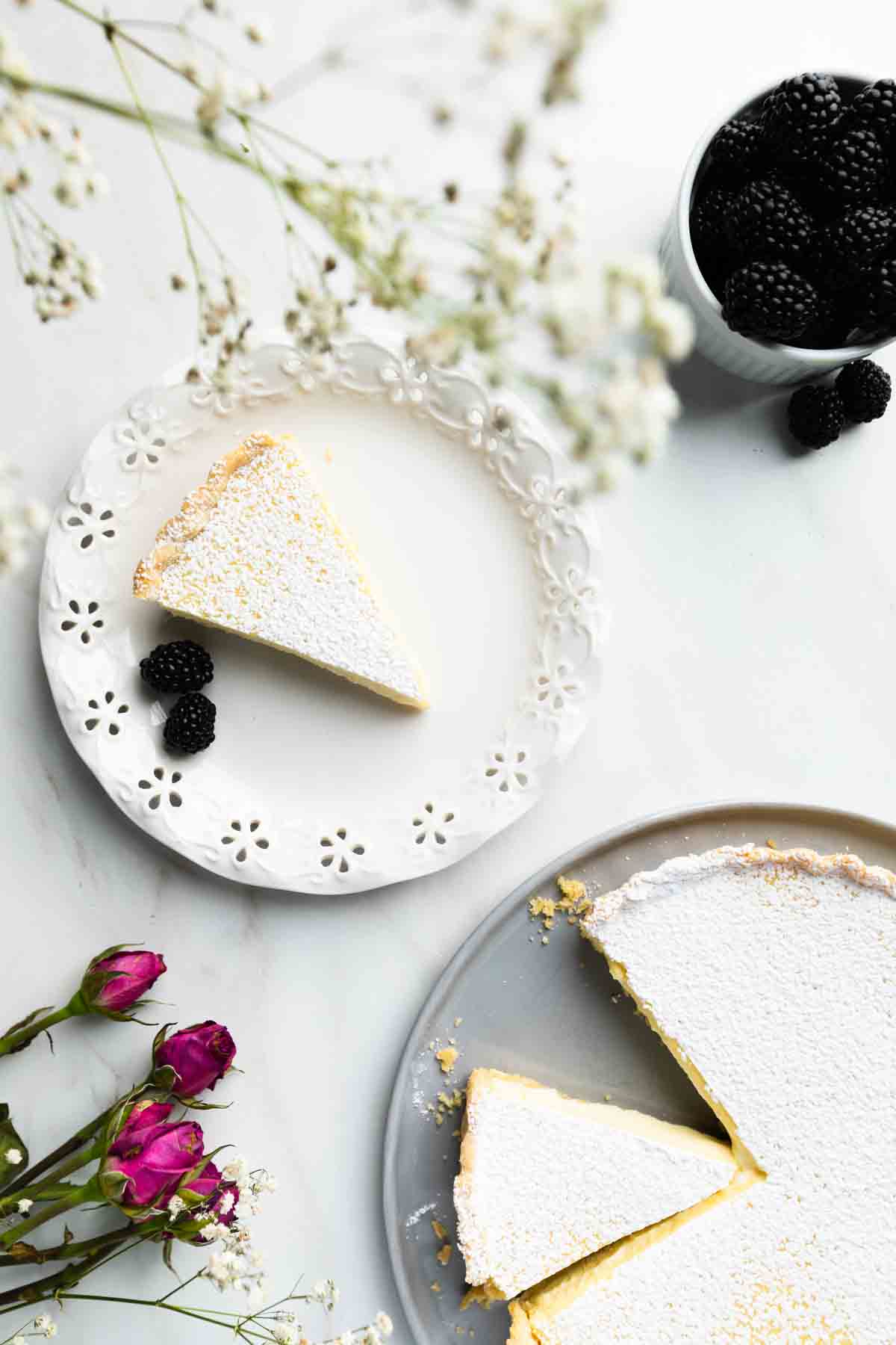 A slice of powdered sugar-topped tart on a white plate with three blackberries, beside a bowl of blackberries and flowers, with the rest of the tart on a gray plate—perfect inspiration for easy pie recipes on National Pie Day.