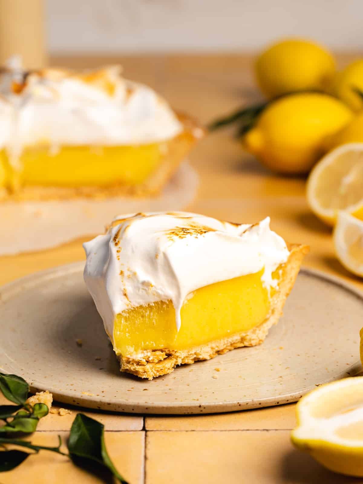 A slice of lemon meringue pie topped with fluffy meringue sits on a plate, with whole and halved lemons in the background—a perfect treat inspired by National Pie Day recipes from the best bloggers.