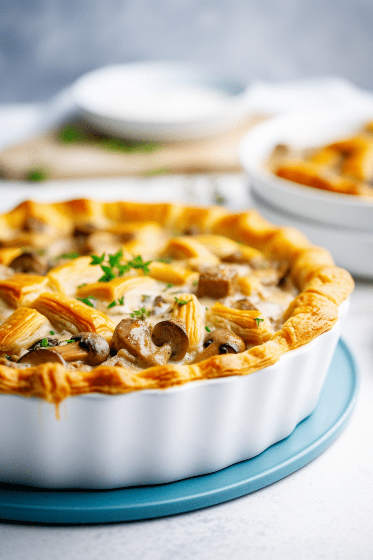 A golden-brown mushroom pie in a white ceramic dish, garnished with fresh herbs, sits on a blue trivet—perfect inspiration for National Pie Day recipes from the best bloggers, with plates visible in the background.