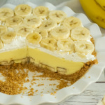 A banana cream pie with a slice removed. The pie features a graham cracker crust, creamy filling, and banana slices on top. Bananas are also visible in the background.