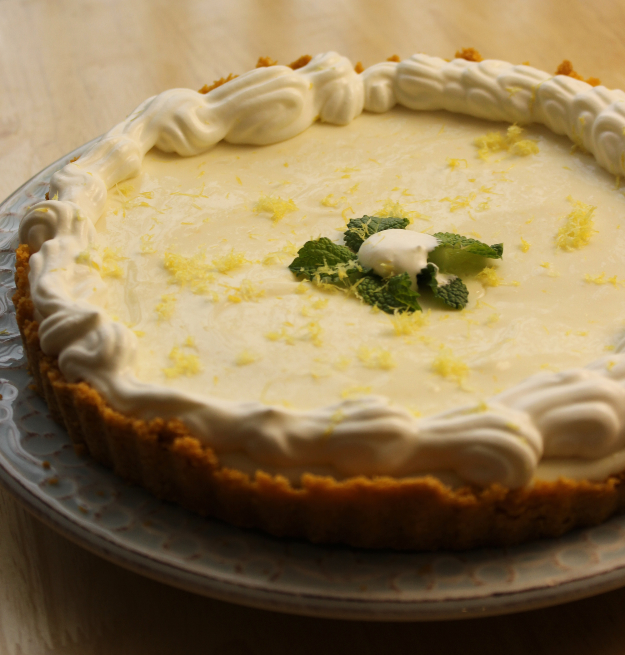 A lemon tart with a graham cracker crust, topped with whipped cream, lemon zest, and mint leaves, on a decorative plate.
