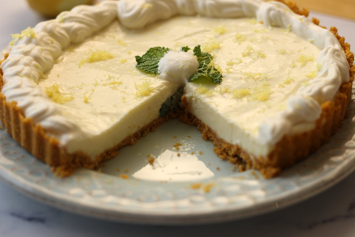 A lemon cream pie with a graham cracker crust, topped with whipped cream, mint leaves, and lemon zest, with one slice removed on a decorative plate.