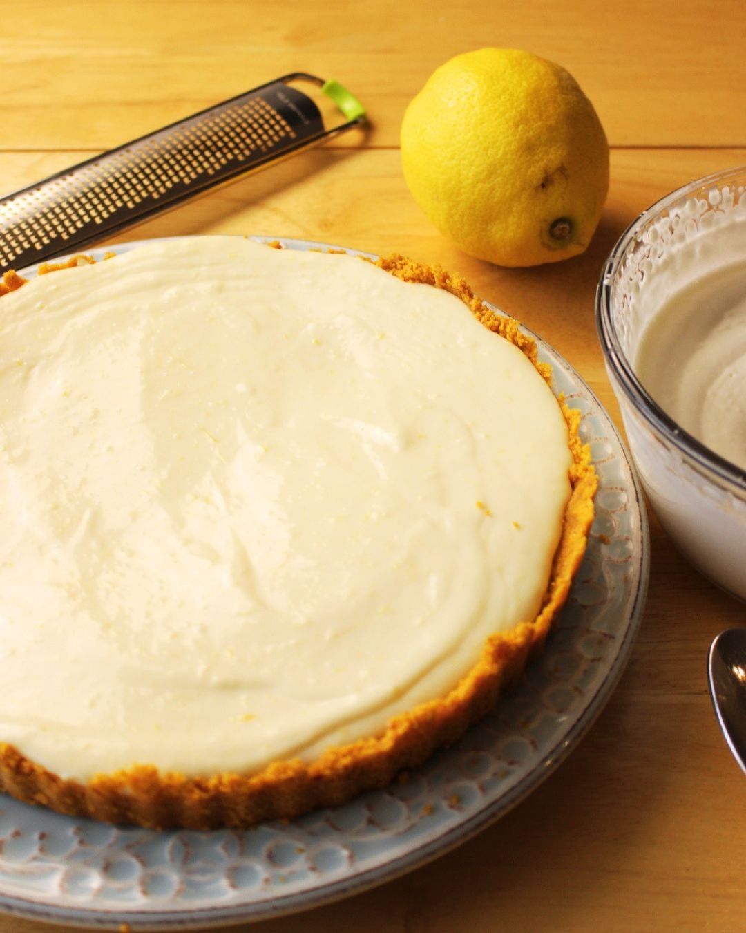 A no bake lemon tart with a creamy filling sits on a plate beside a fresh lemon, a zester, and a bowl of cream on a wooden table—an easy lemon dessert perfect for any occasion.