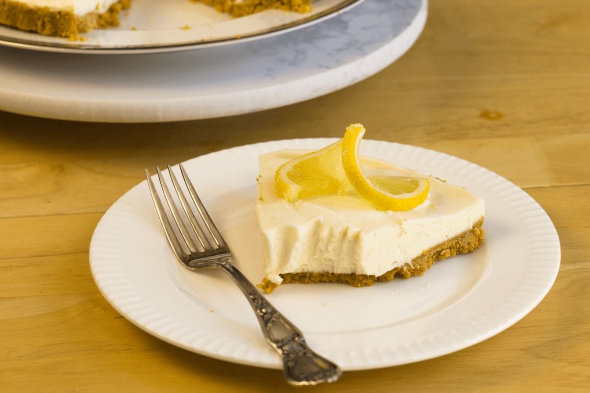 A slice of lemon cheesecake with a graham cracker crust is served on a white plate with a fork, garnished with lemon slices—perfect for those seeking an easy lemon tart recipe or inspiration for a no bake lemon tart.